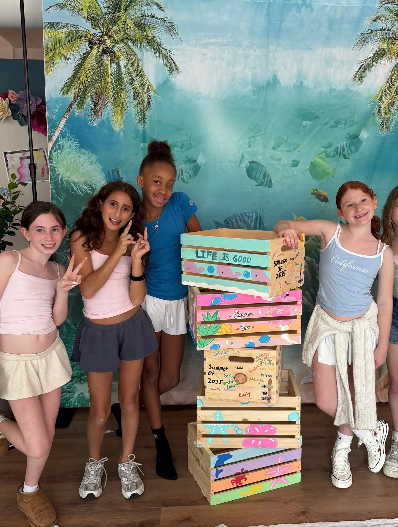 Friends stand beside a tower of crates they painted at a custom party, with an ocean backdrop.